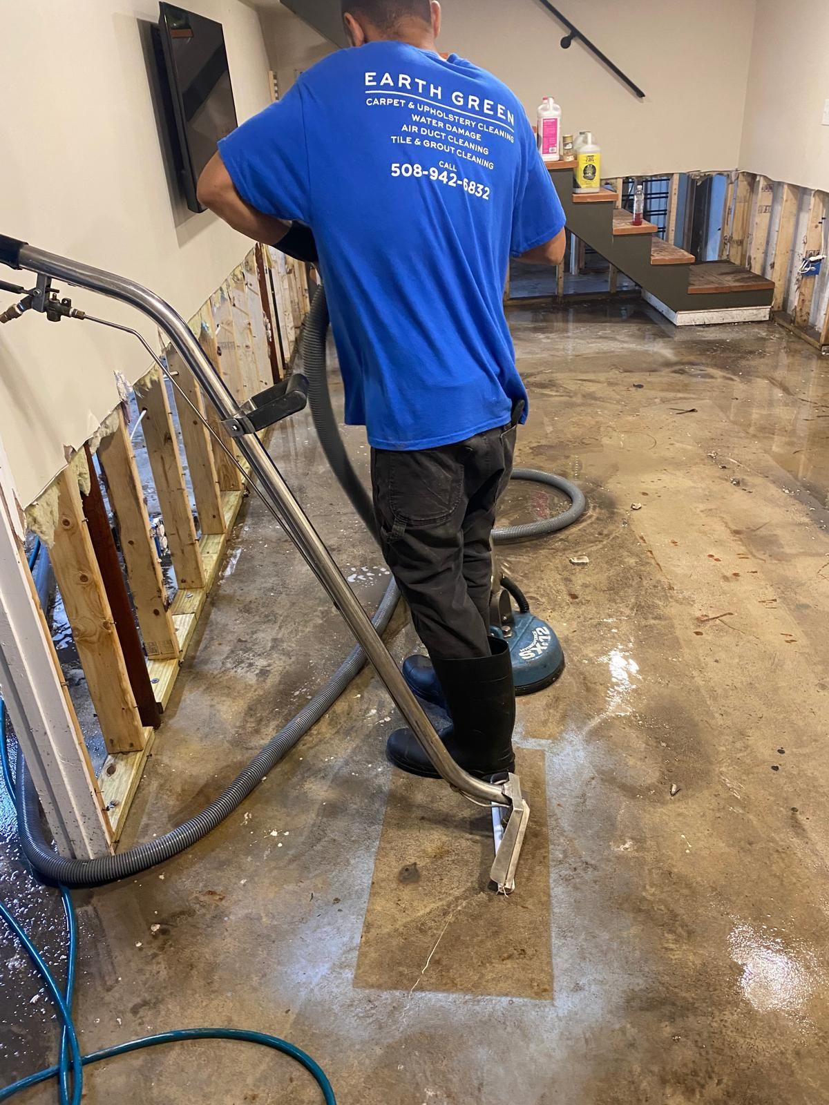 A worker in a blue uniform uses an industrial vacuum on a wet concrete floor in a room with partially dismantled walls.