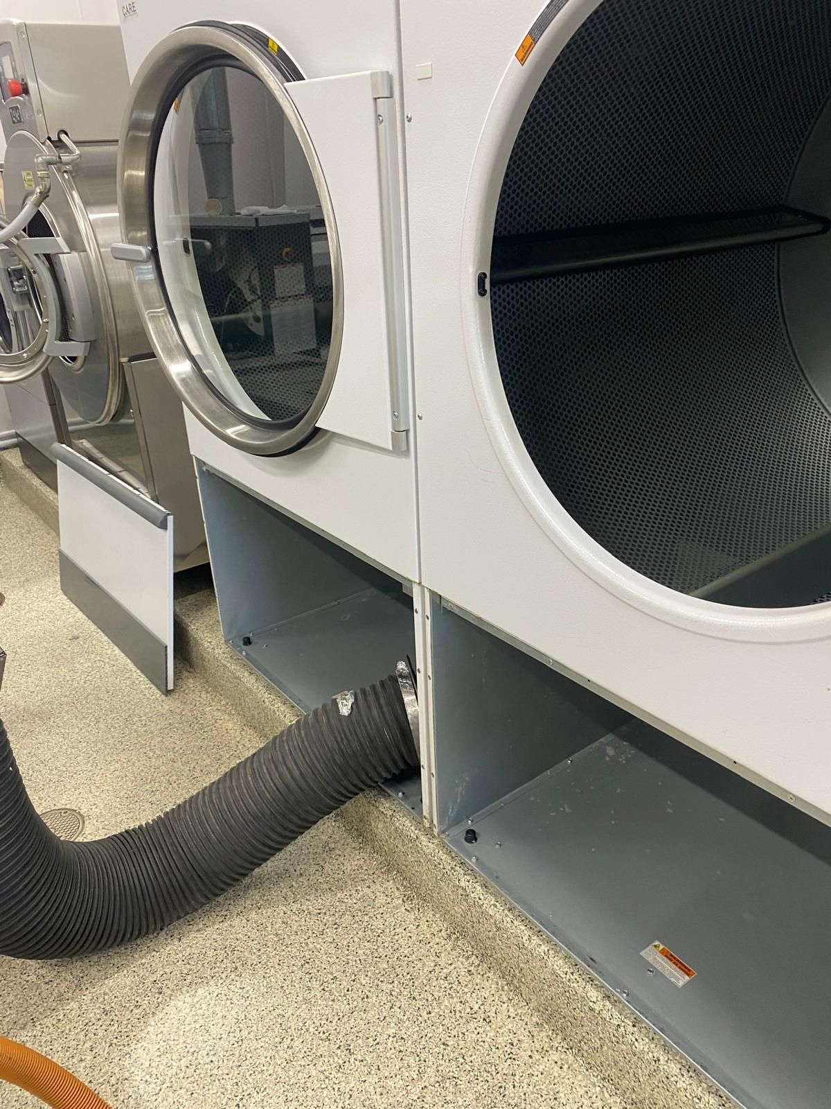 Two white commercial laundry dryers side-by-side in a facility, with a flexible black vent hose connected to one base.