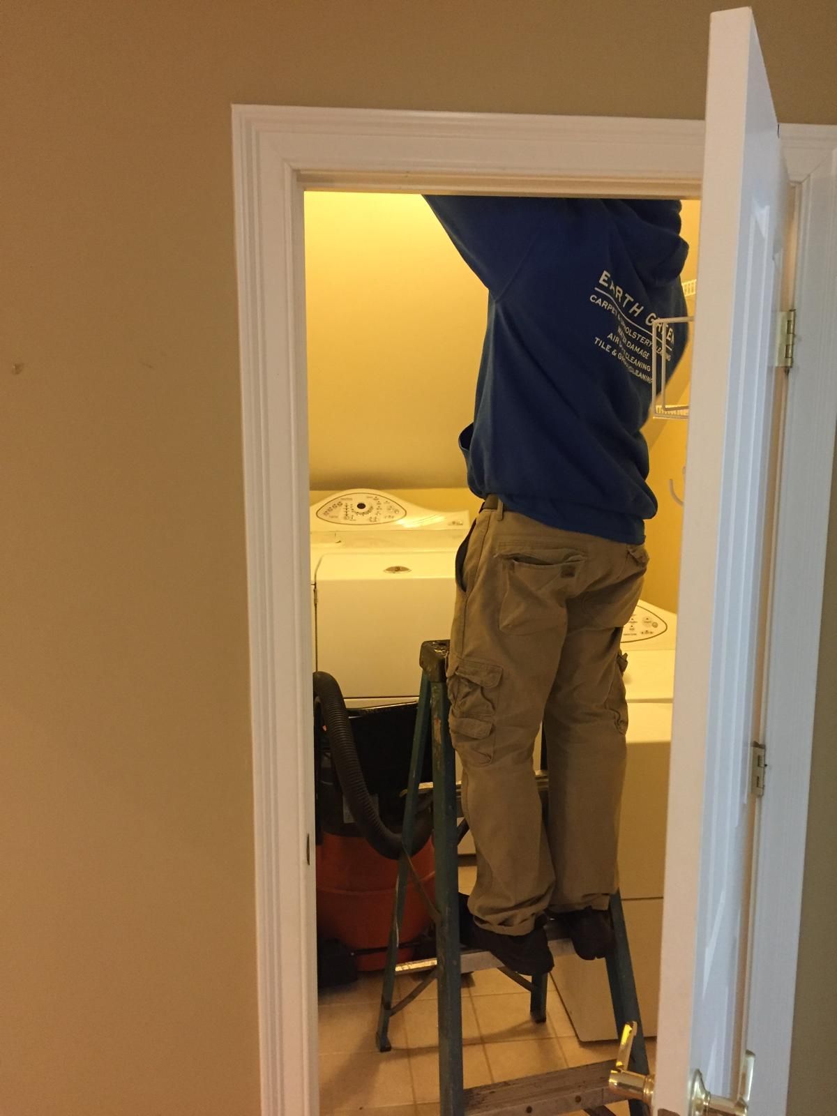 A person in a blue hoodie and tan cargo pants stands on a stepladder, working on the ceiling inside a small closet.