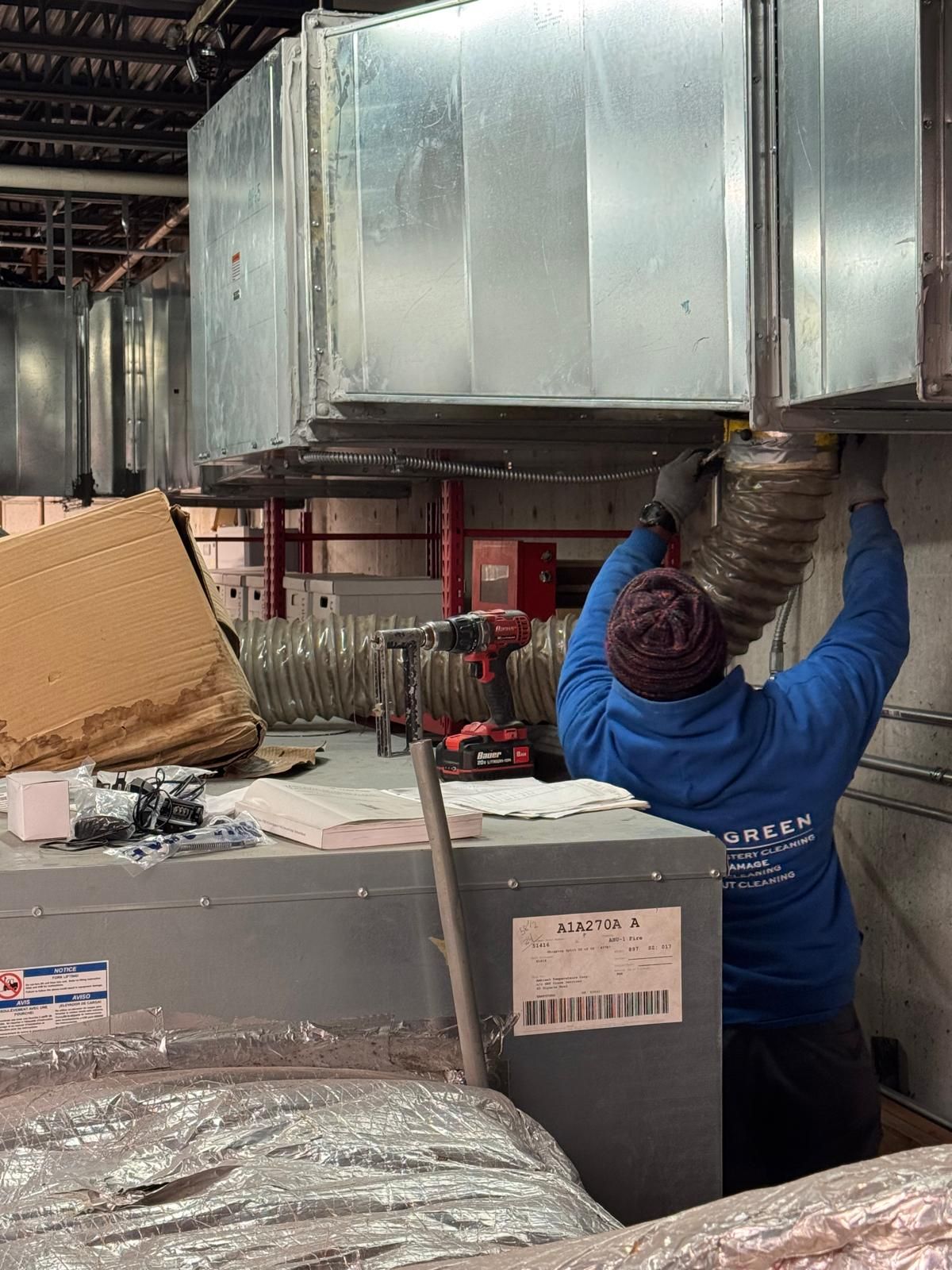 A person in a blue hoodie works on ceiling HVAC ductwork, holding a flexible vent hose near a metal box in an industrial site.