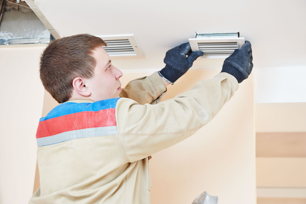 A technician wearing work clothes and gloves installs or adjusts a white ceiling ventilation vent.