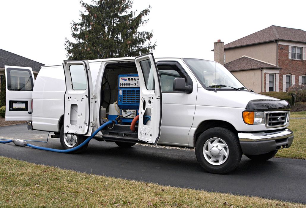 White utility van with open side and rear doors, parked on a driveway with carpet cleaning equipment visible inside.