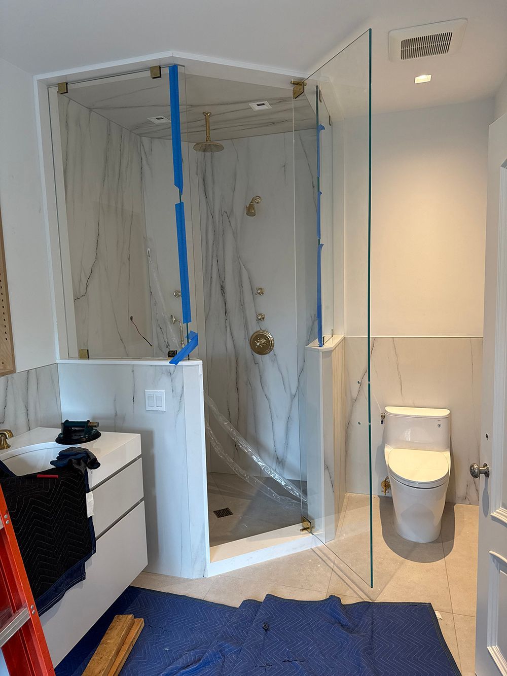 Bathroom under renovation with shower area masked in plastic and blue tape, glass partition, and toilet visible