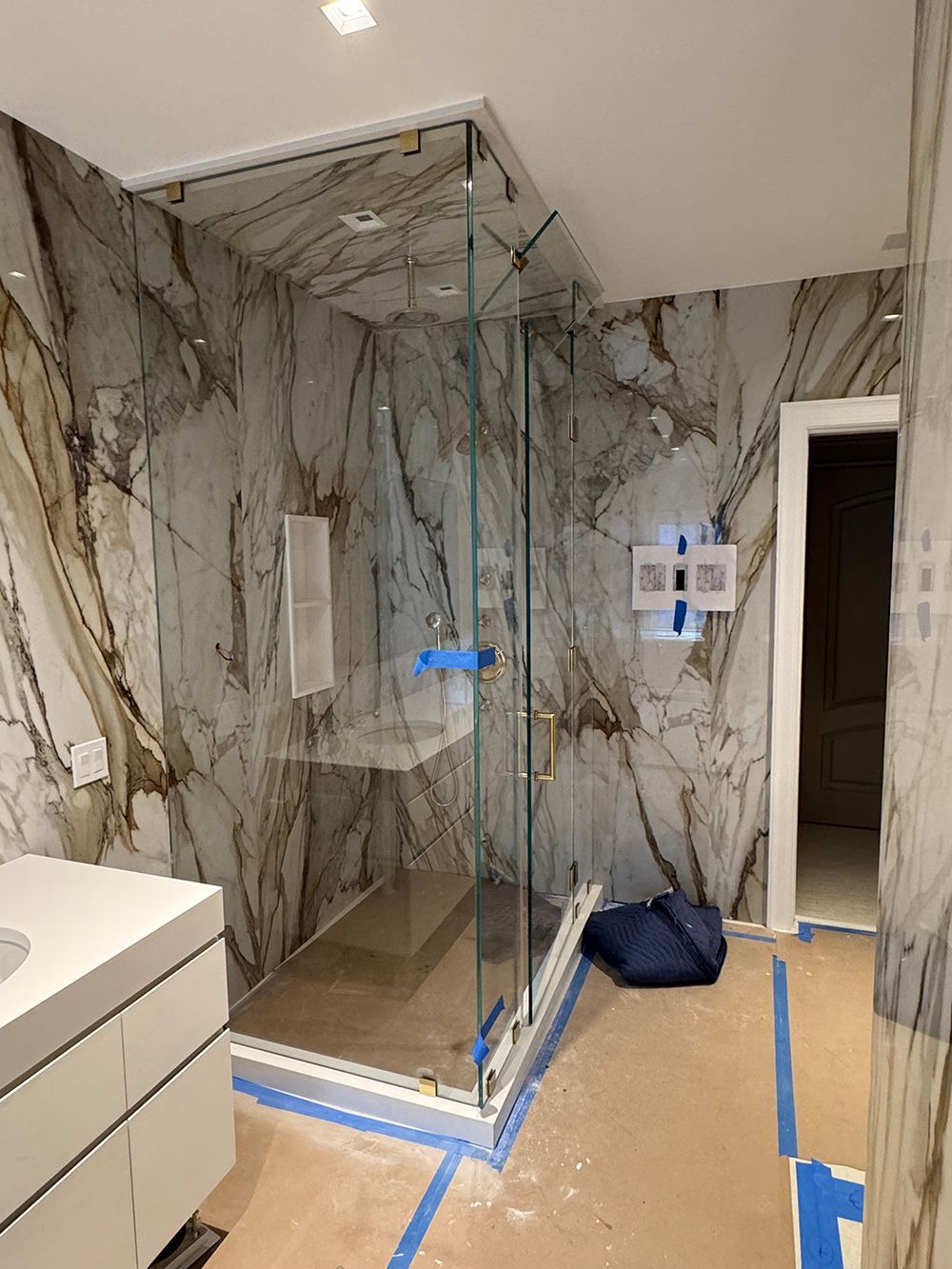 Modern bathroom shower with marble tile, glass enclosure, and vanity at left; blue tape on floor.