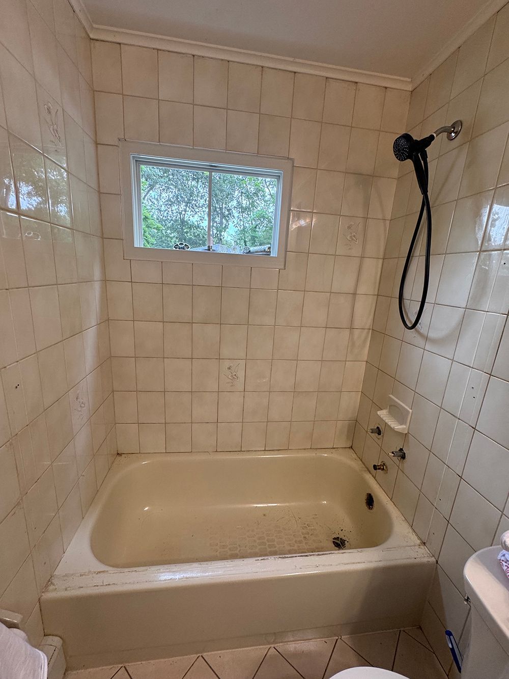 Small tiled bathroom with a cream bathtub, window above, and a handheld shower on the right.