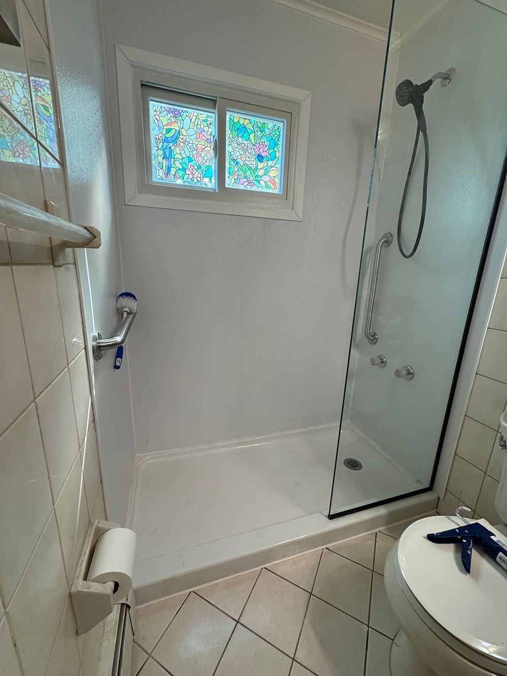 Small white tiled bathroom with shower stall, frosted window, and sink in the foreground.