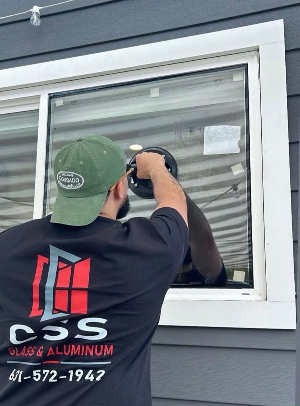 Worker in black shirt and green cap installing a window on a gray house exterior.