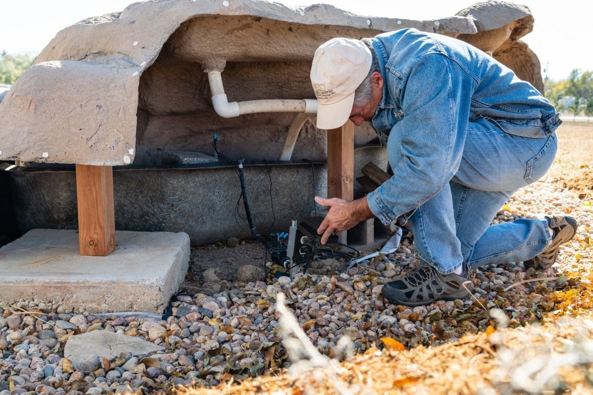 A man is kneeling down in the dirt looking at a pipe.