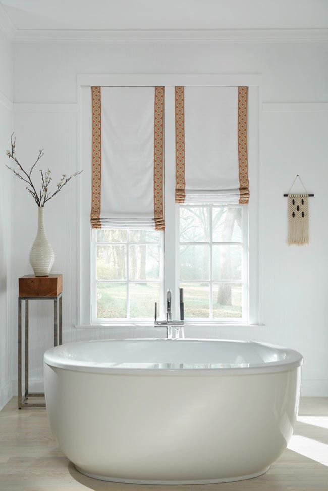 A bathroom with a tub and a window with roman shades.