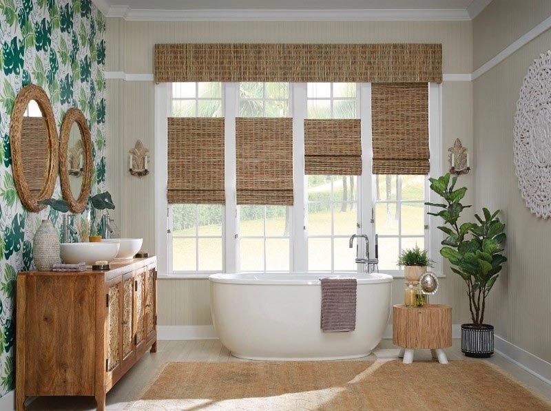 A bathroom with a bathtub , sink , mirrors and wicker blinds.
