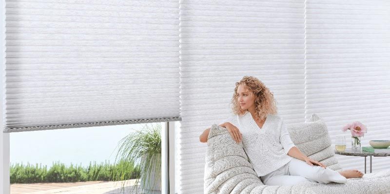 A woman is sitting on a couch in front of a window with white blinds.