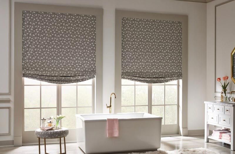 A bathroom with a bathtub and two windows with roman shades.