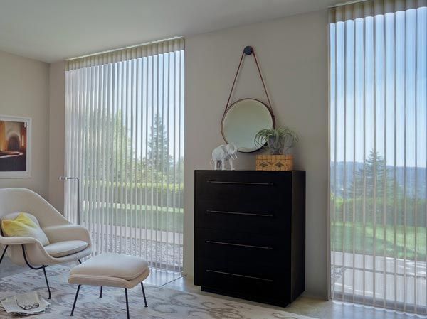 A living room with vertical blinds , a chair , a dresser and a mirror.