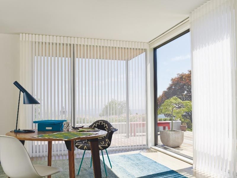 A room with a table and chairs and a window with vertical blinds.
