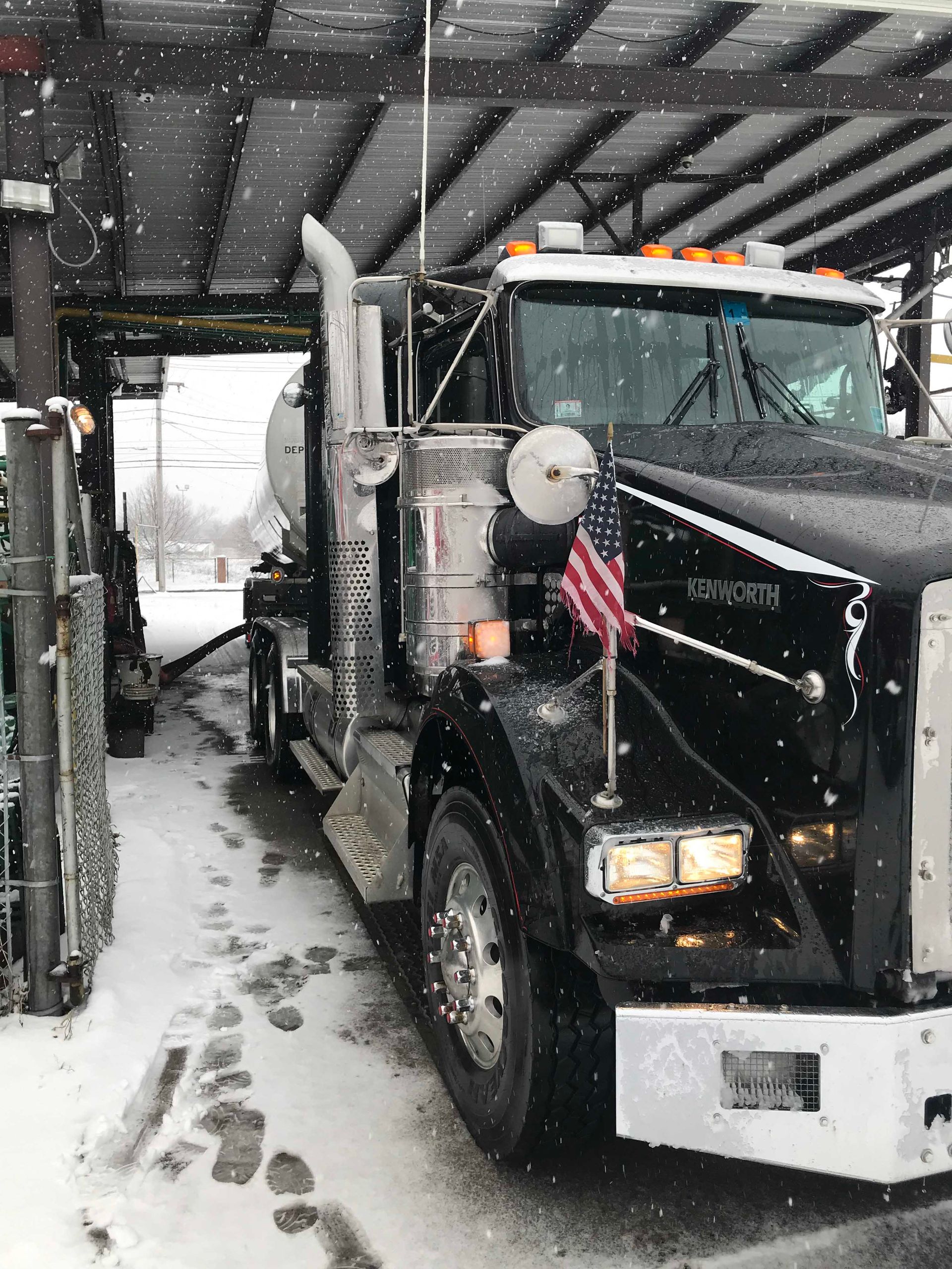 Black semi truck with trailer driving through snowy underpass, American flag on the side.