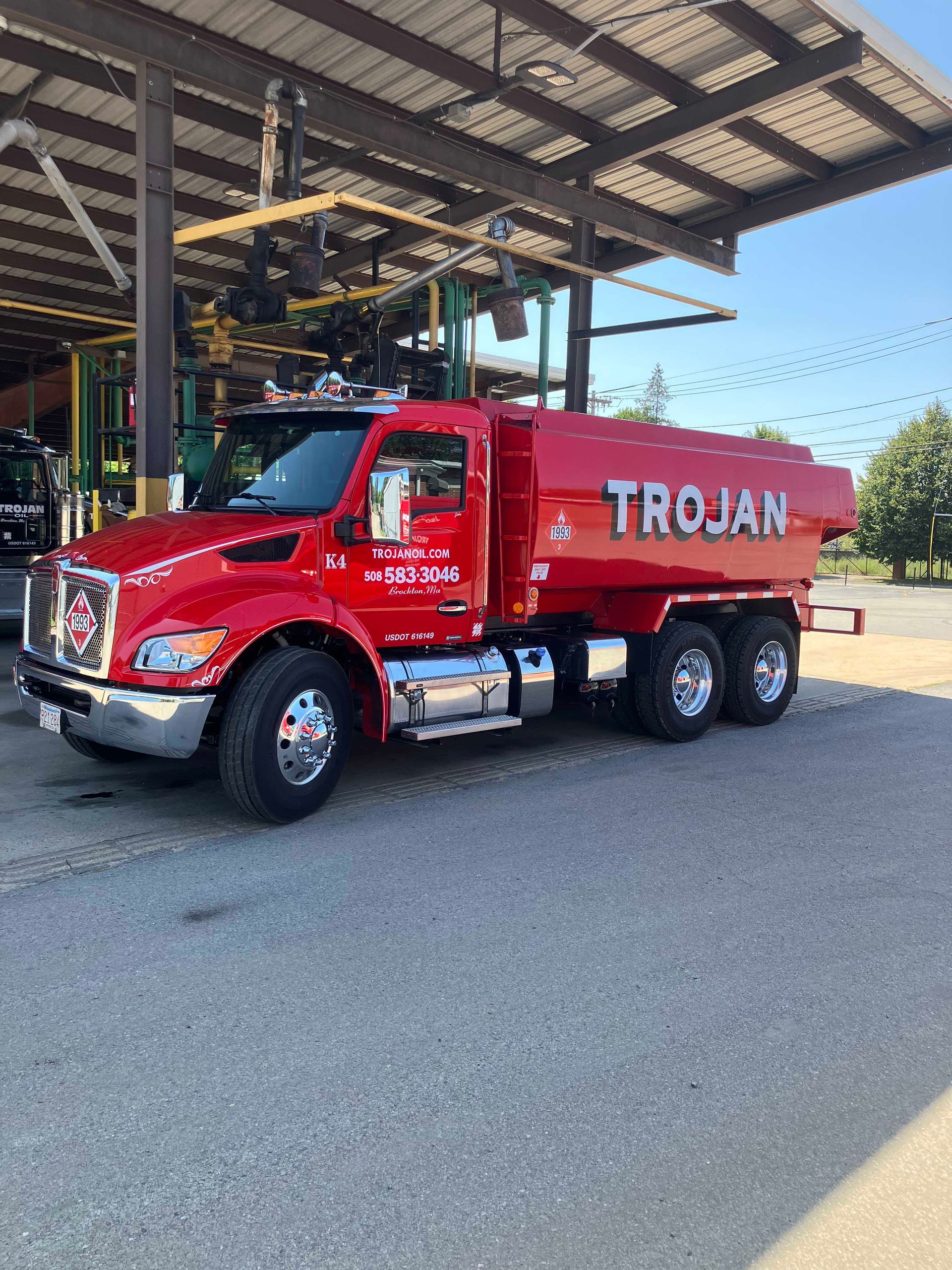 Red TROJAN tanker truck parked under a metal canopy in an industrial yard.