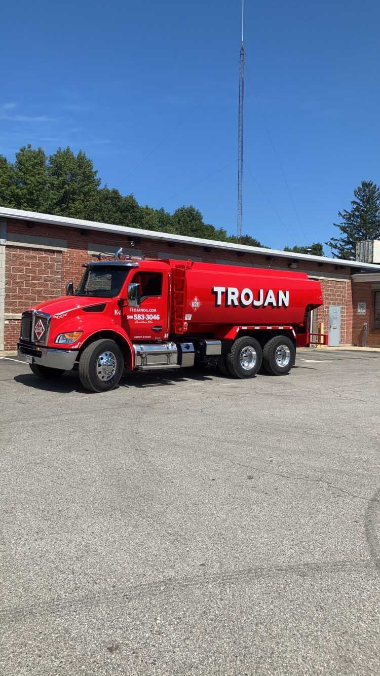 Red Trojan tanker truck parked on gravel beside a brick building under a clear blue sky