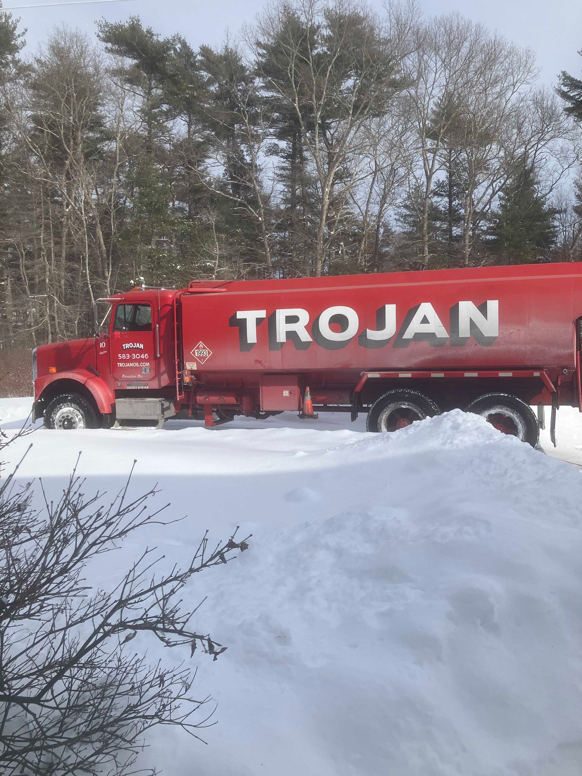 Red Trojan tanker truck driving through snow on a road with trees in the background.
