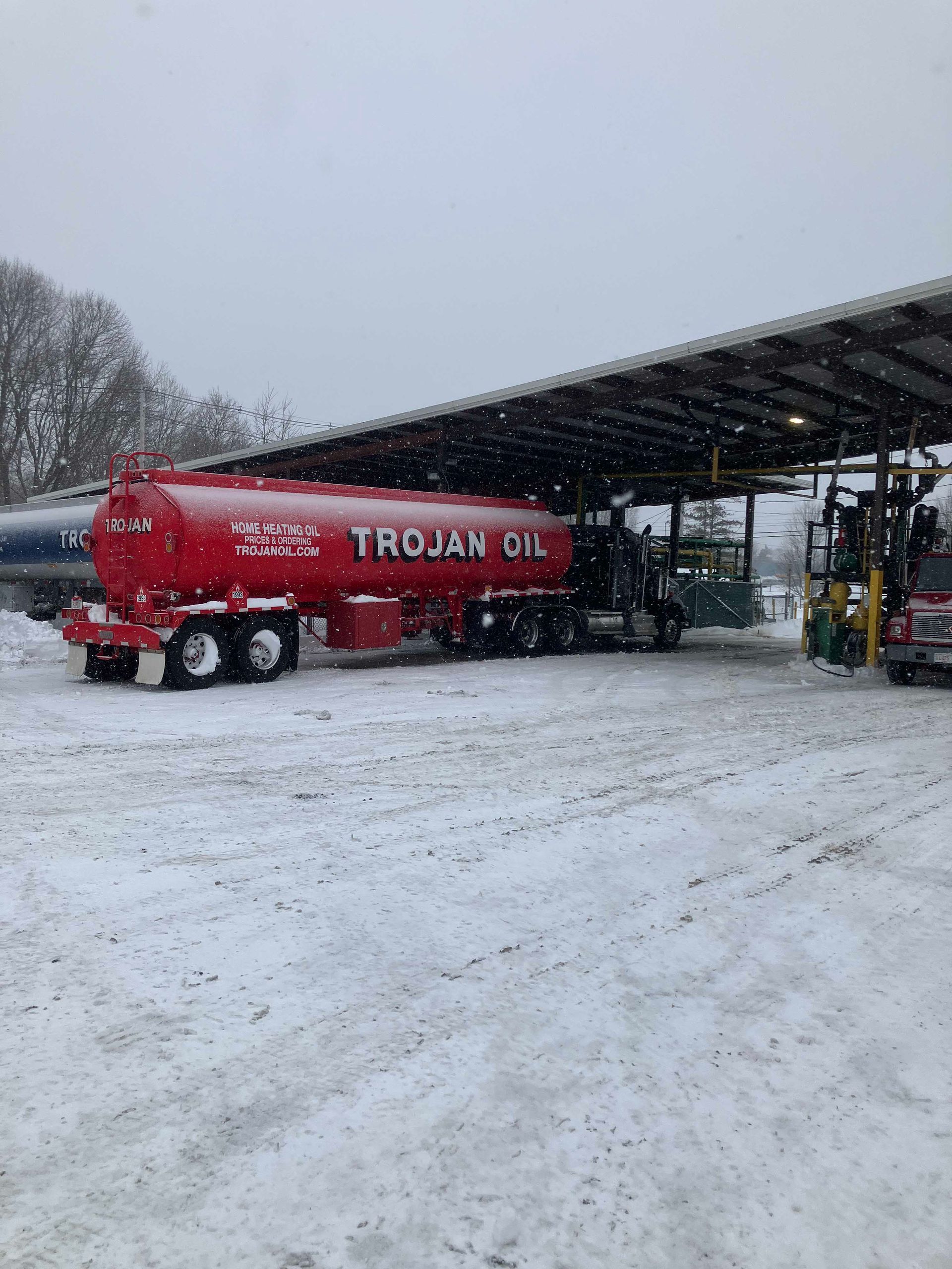 Red Trojan Oil tanker truck parked in a snowy industrial yard under a canopy.