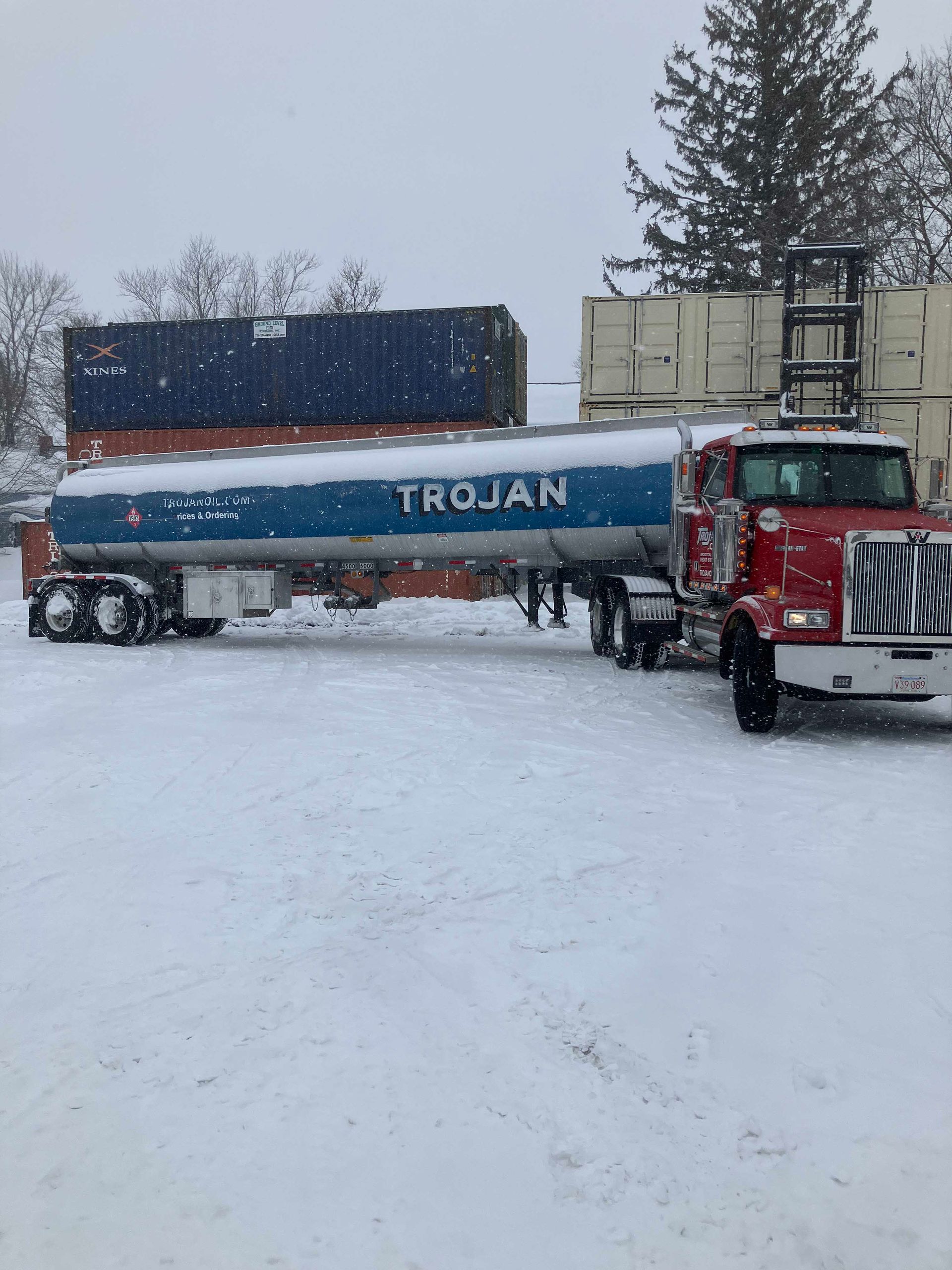 Red and white semi-truck hauling a blue TROJAN container through a snowy lot