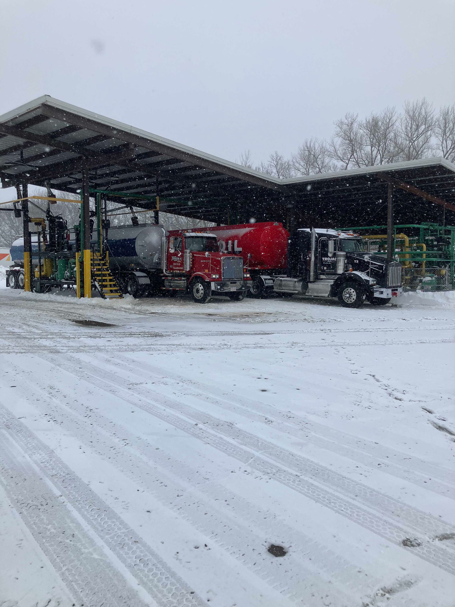 Snow-covered truck yard with a red tanker under a large metal canopy in falling snow