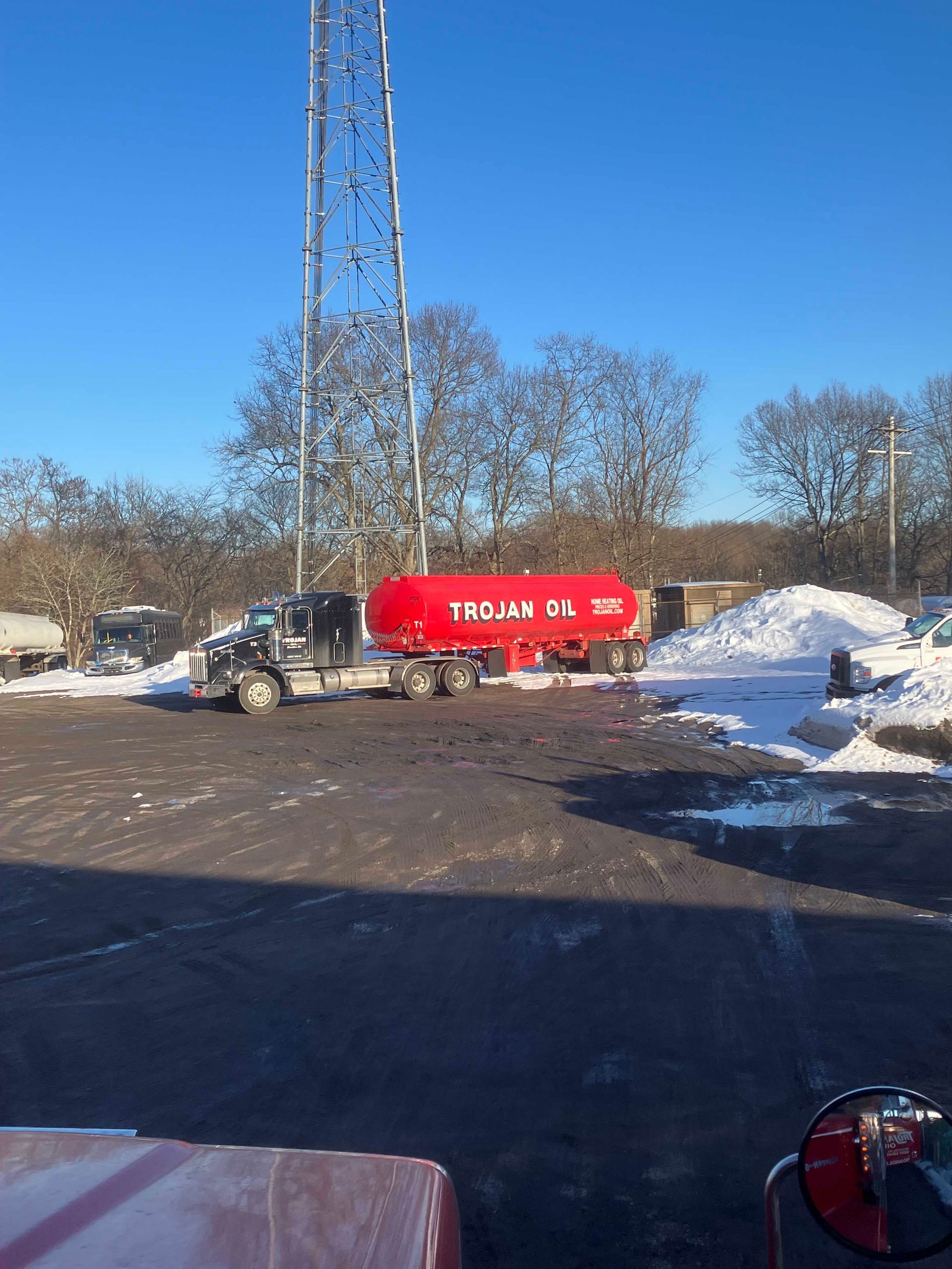 Red tanker truck parked in a snowy lot beside a tall metal tower under a clear blue sky.