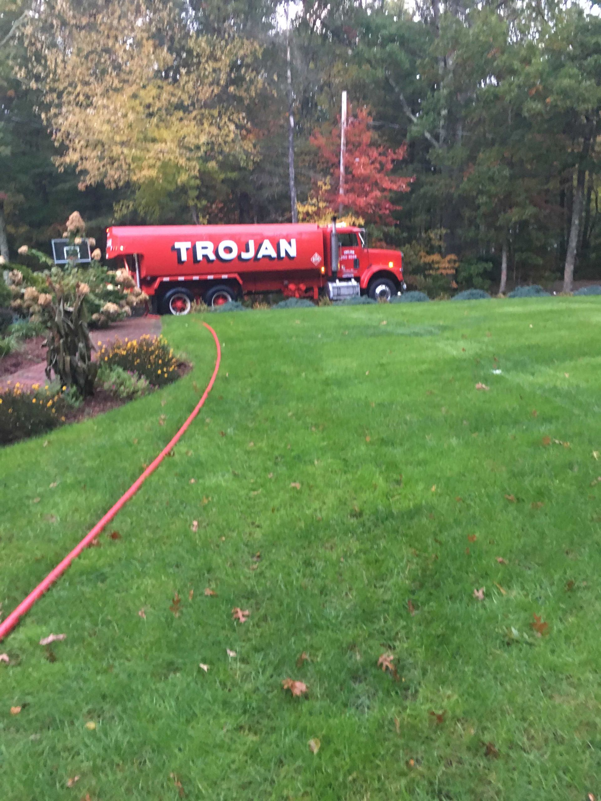 Red TROJAN water truck parked on a grassy lawn near trees and a hose in autumn.