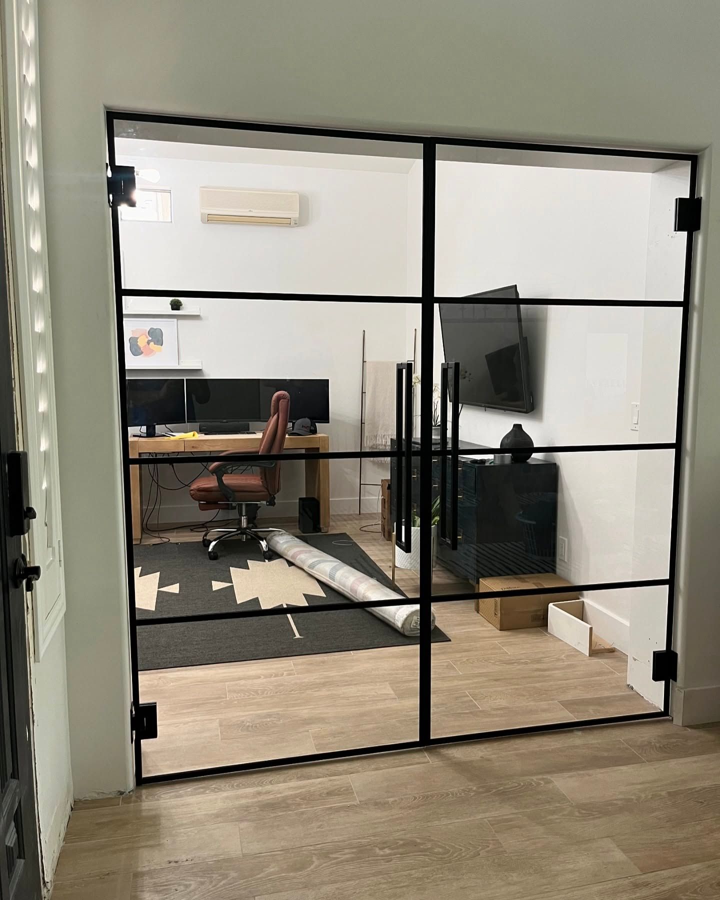 Home office seen through black-framed glass doors with a desk, chair, TV, and rug.