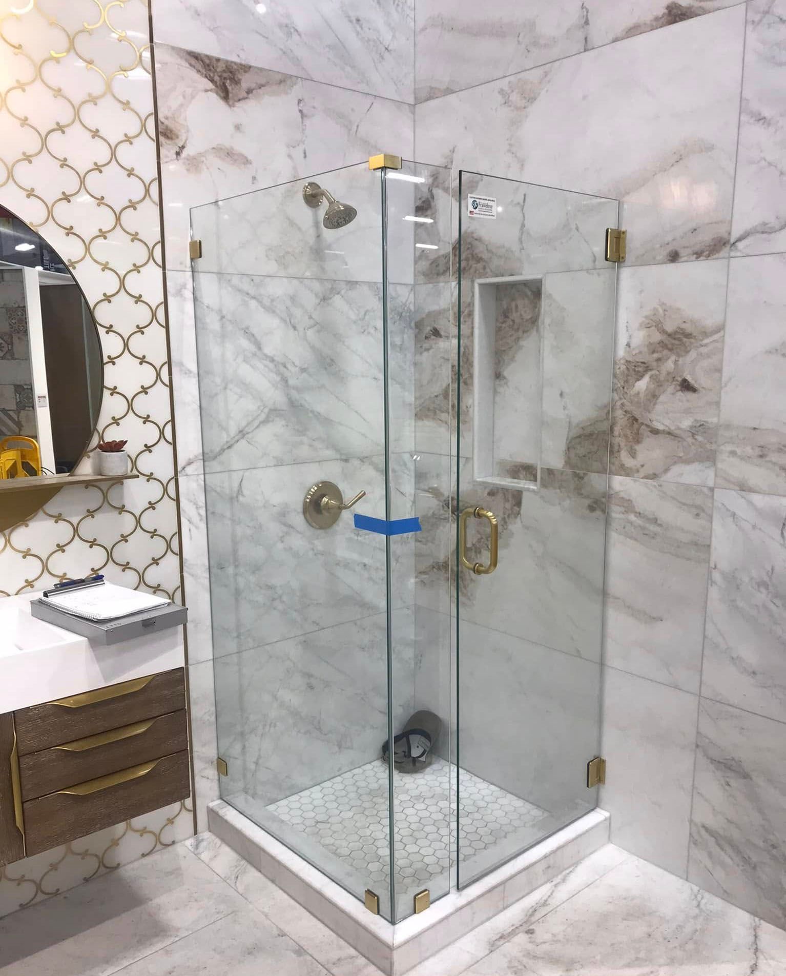 Glass shower enclosure in a marble-tiled bathroom with gold fixtures and a vanity mirror on the left.