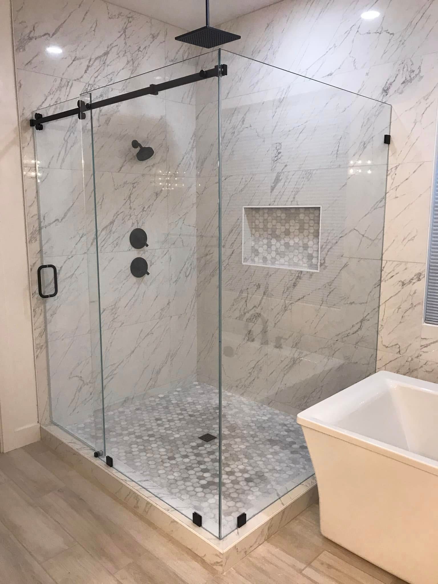 Modern walk-in shower with glass enclosure, marble tile walls, black fixtures, and a white bathtub nearby.