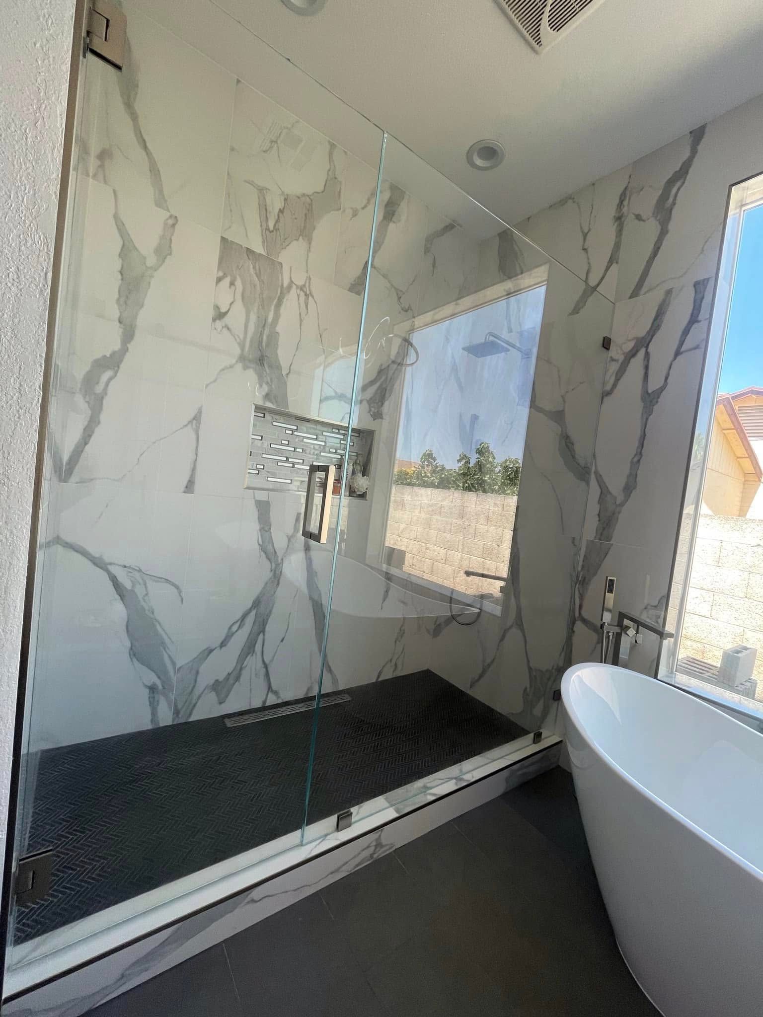 Modern bathroom shower with marble-pattern walls, black floor, and a white freestanding tub beside a window.