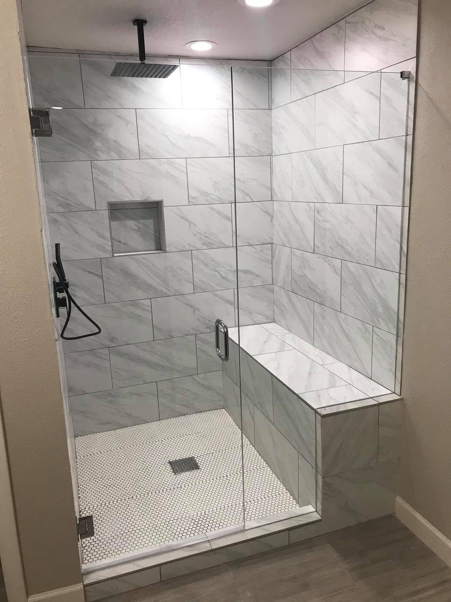 Walk-in tiled shower with glass door, bench, and rainfall showerhead on a marble-look wall.