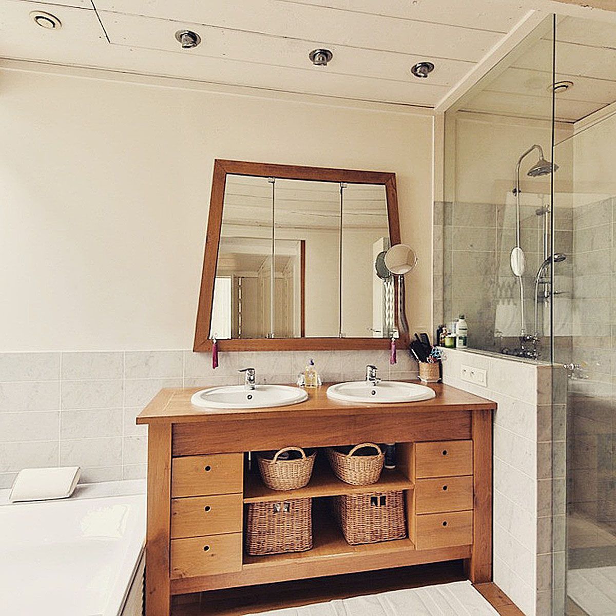 Bathroom with wooden vanity, sink, mirror, and glass shower beside a bathtub; white tile walls.