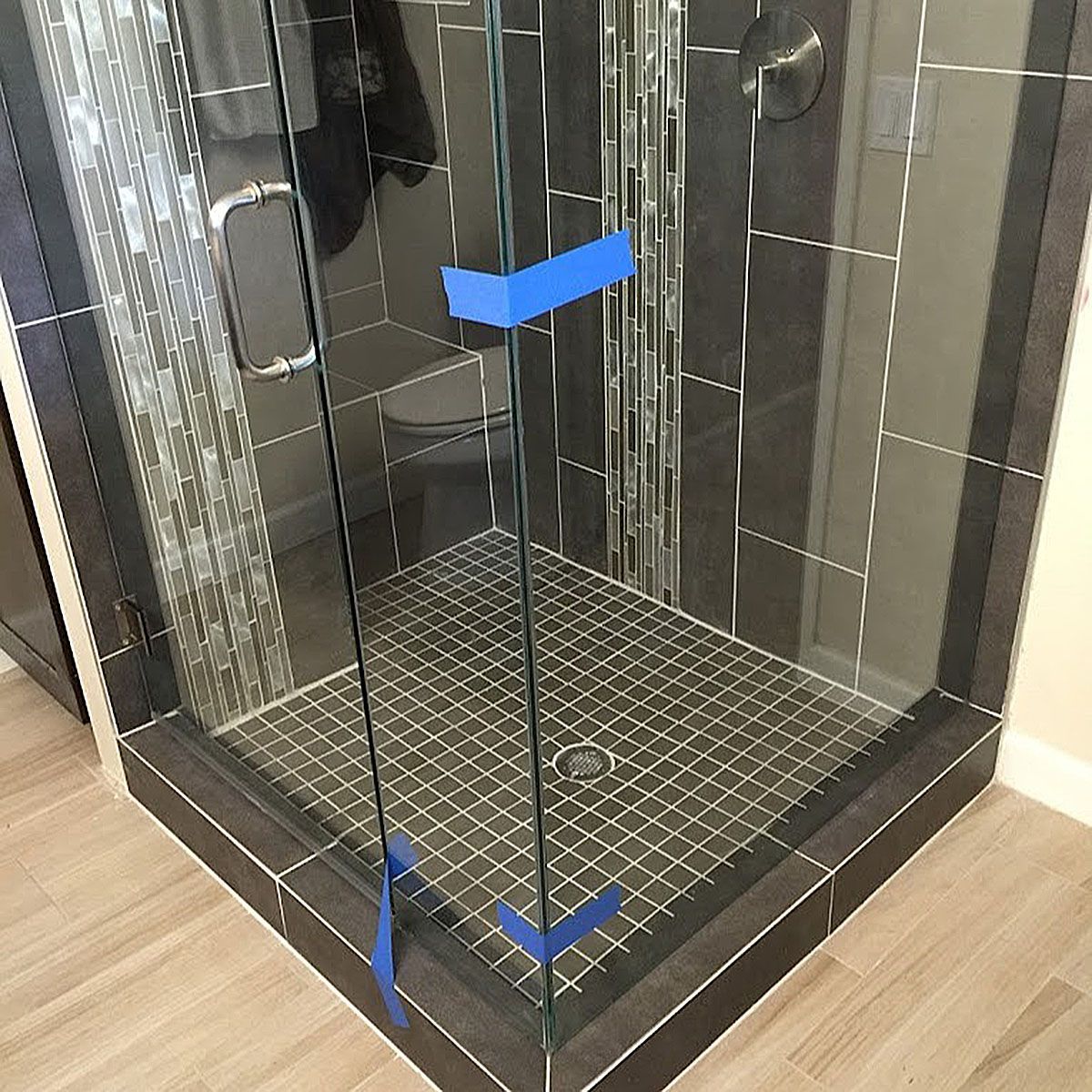 Accessible shower stall with blue grab bars, black tile walls, and a diamond-pattern floor near wooden flooring.