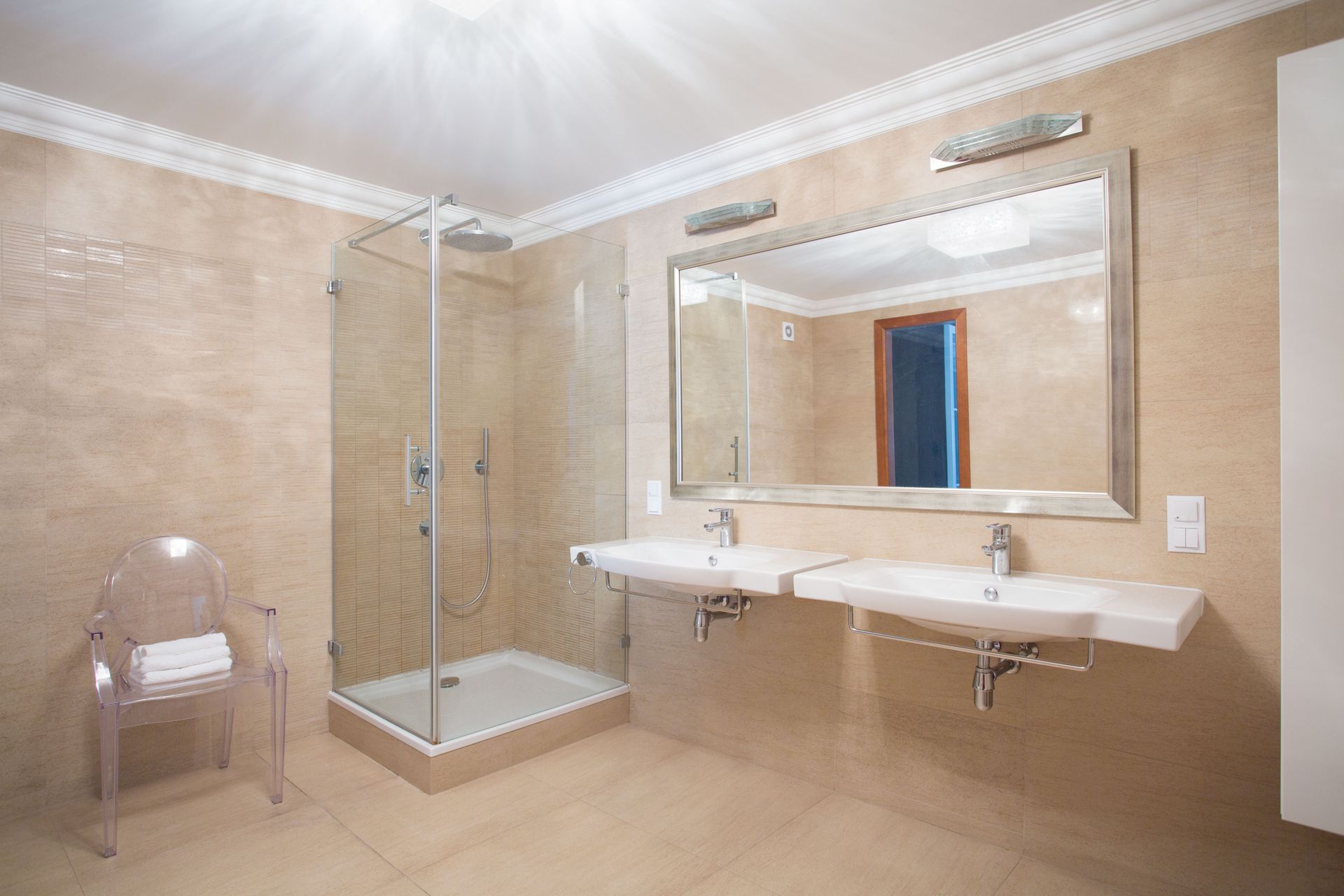 Bright beige hotel bathroom with glass shower, double sink, large mirror, and chair.