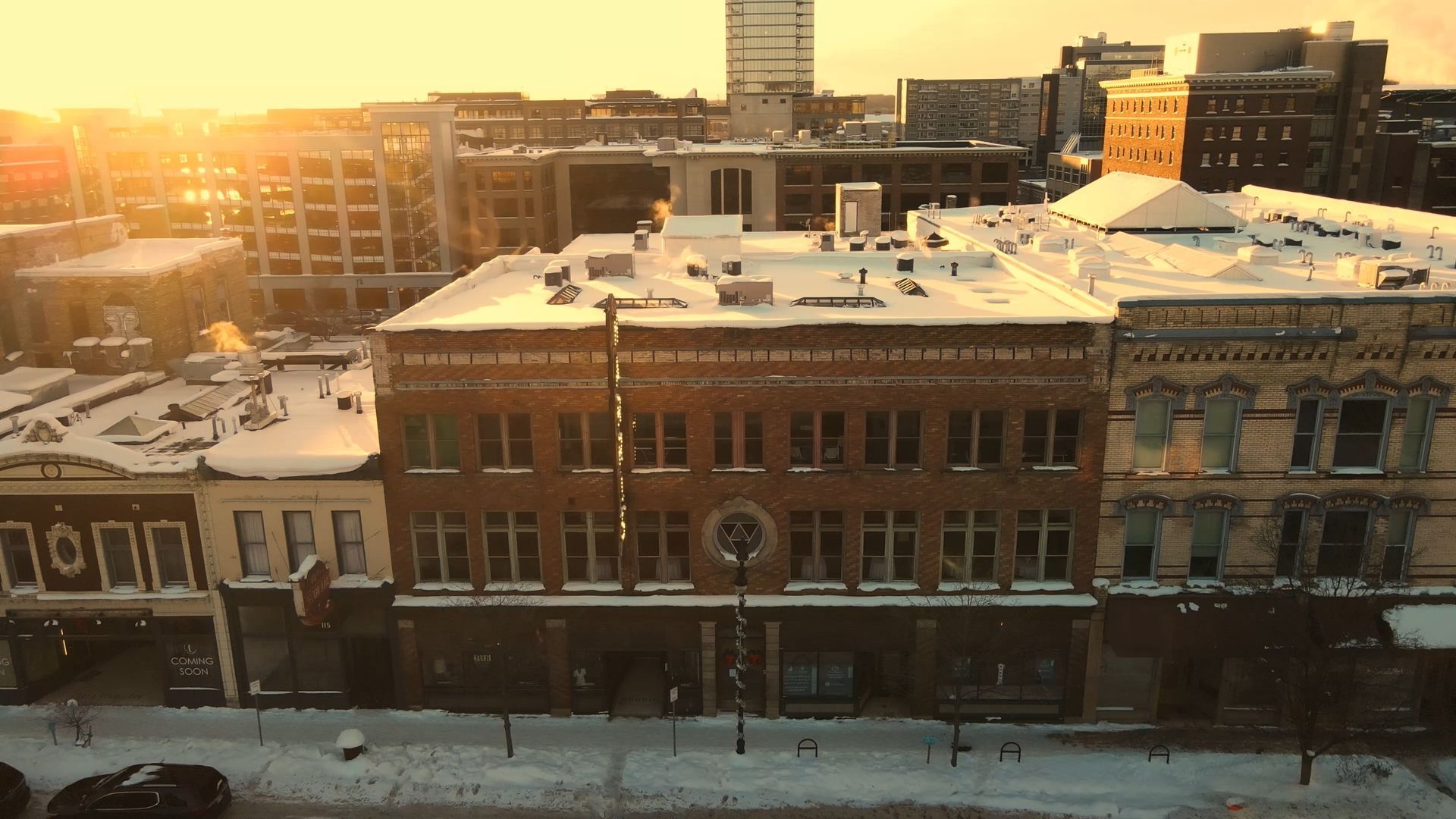 Snowy city street with brick buildings at sunset, warm sunlight glowing over rooftops.