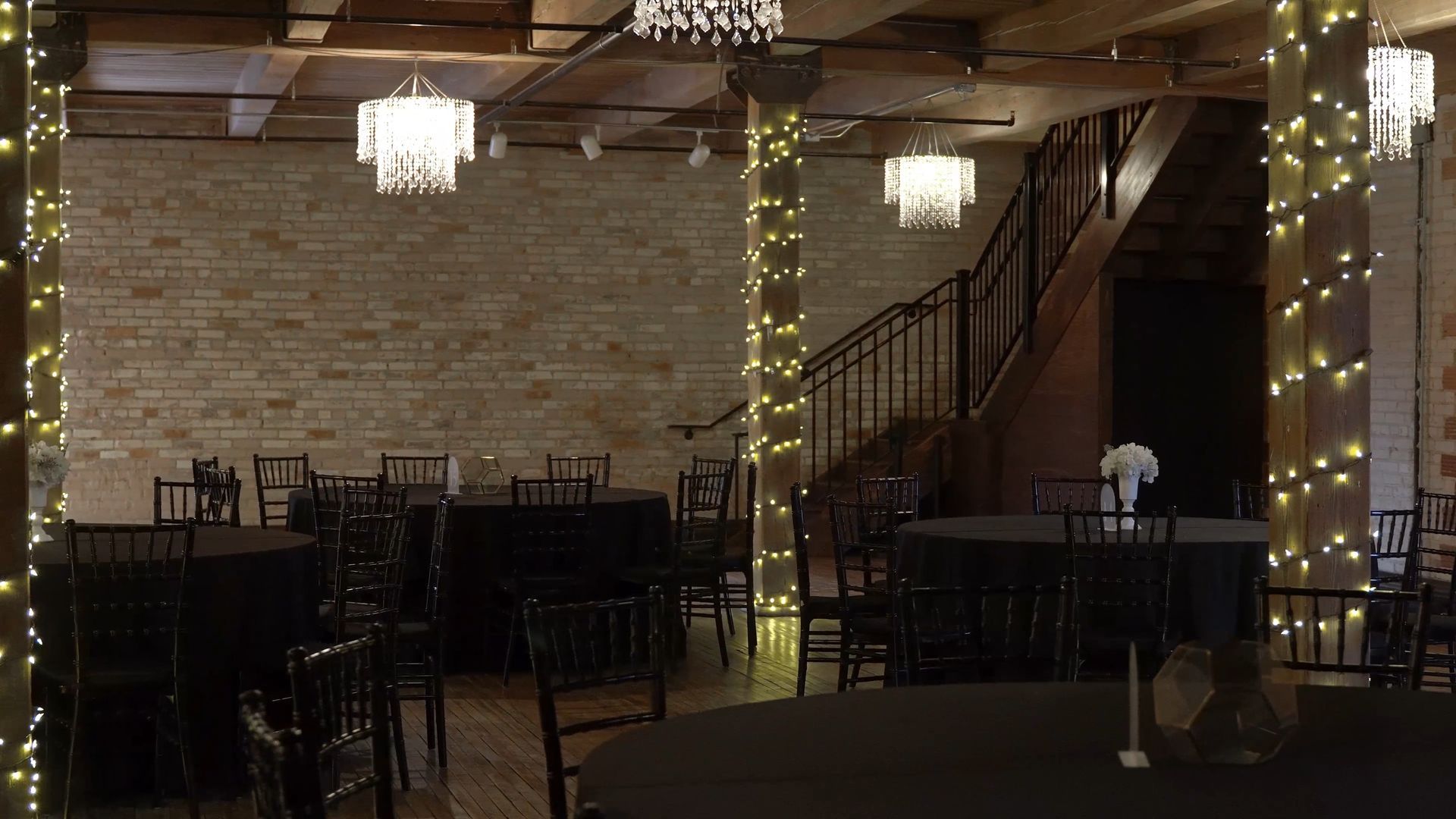 Rustic event hall with stone walls, string lights, chandeliers, and empty tables and chairs.