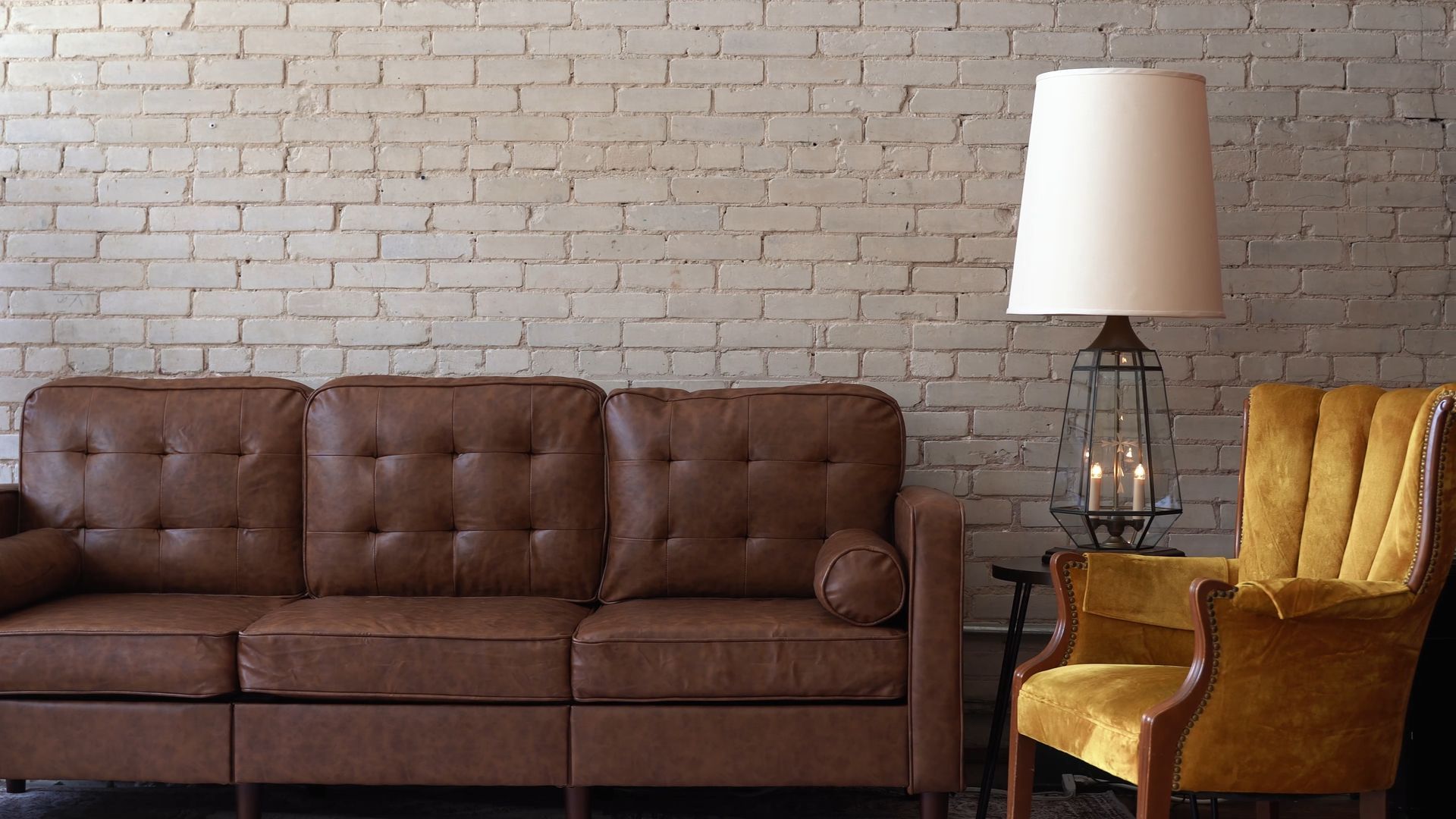Brown leather sofa and gold armchair beside a floor lamp against a white brick wall.