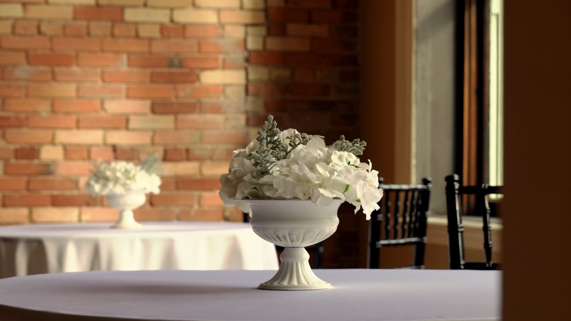 White floral arrangement in a pedestal vase on a table, with a brick wall and chairs in the background.