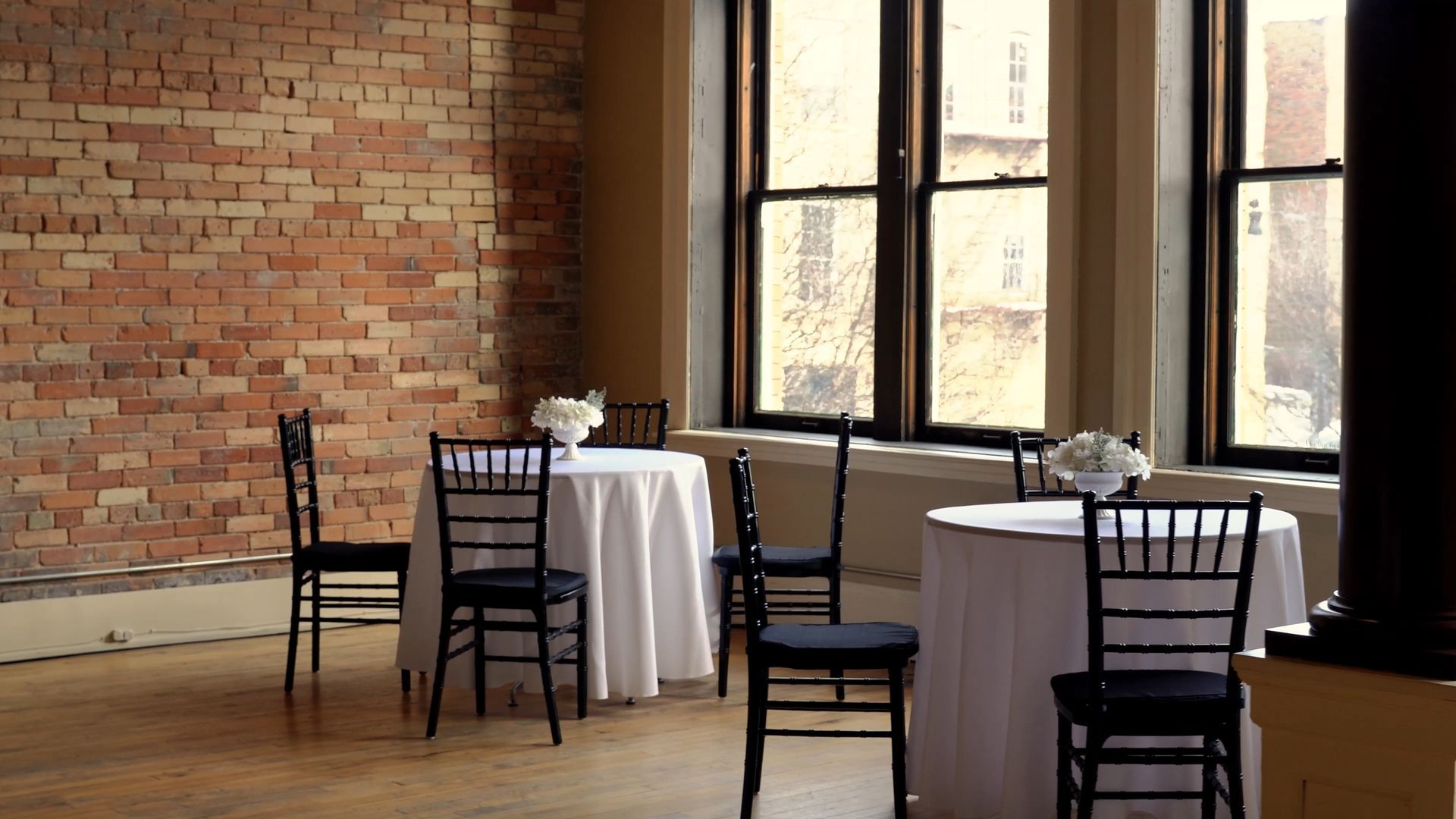 Empty dining area with white tablecloths, black chairs, exposed brick wall, and tall windows.