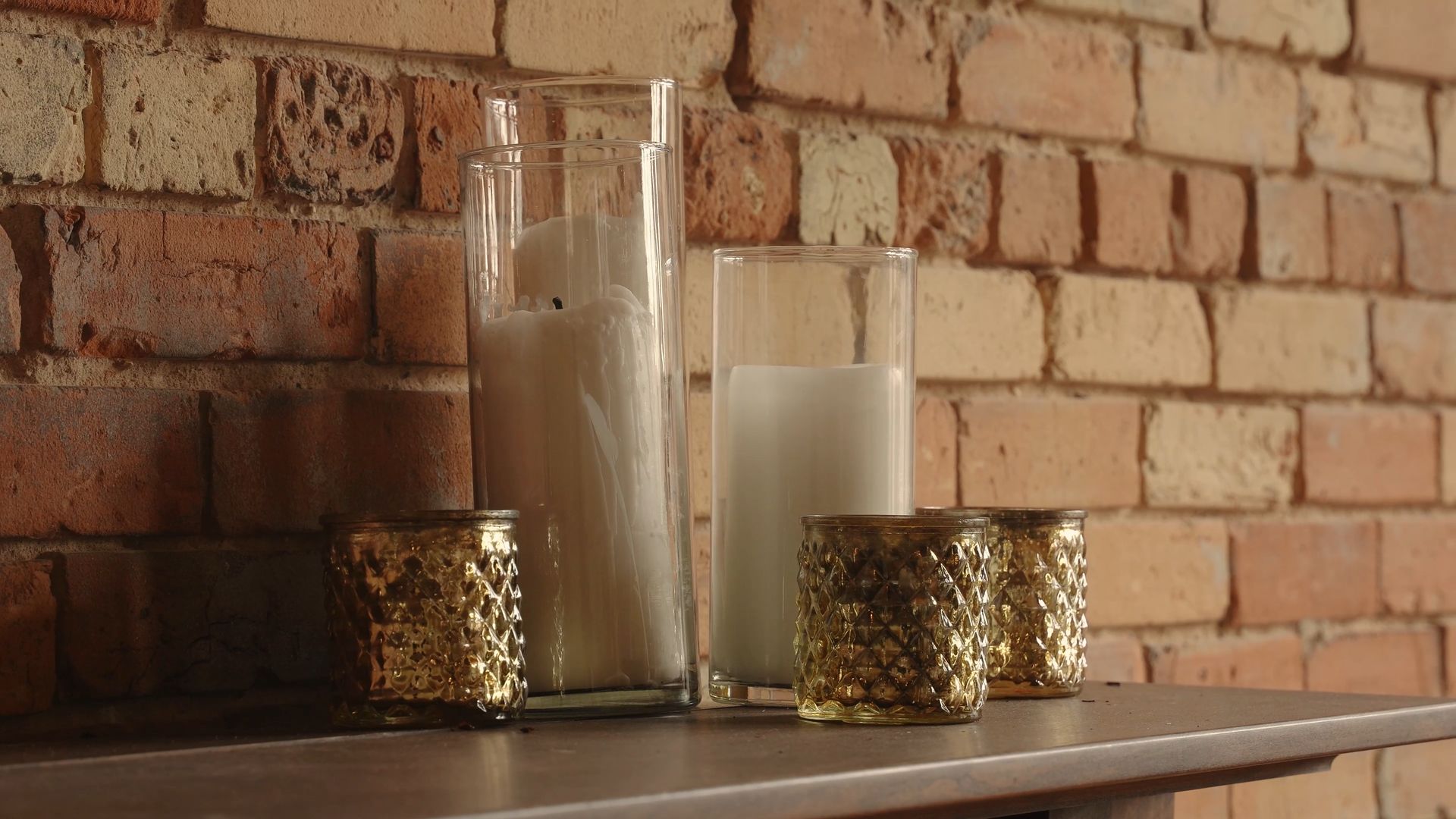 Two white candles in glass holders on a wooden shelf against a brick wall.