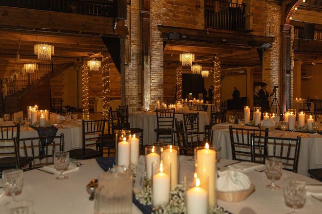 Candles on banquet tables in a warmly lit rustic event hall with exposed brick and hanging lights.
