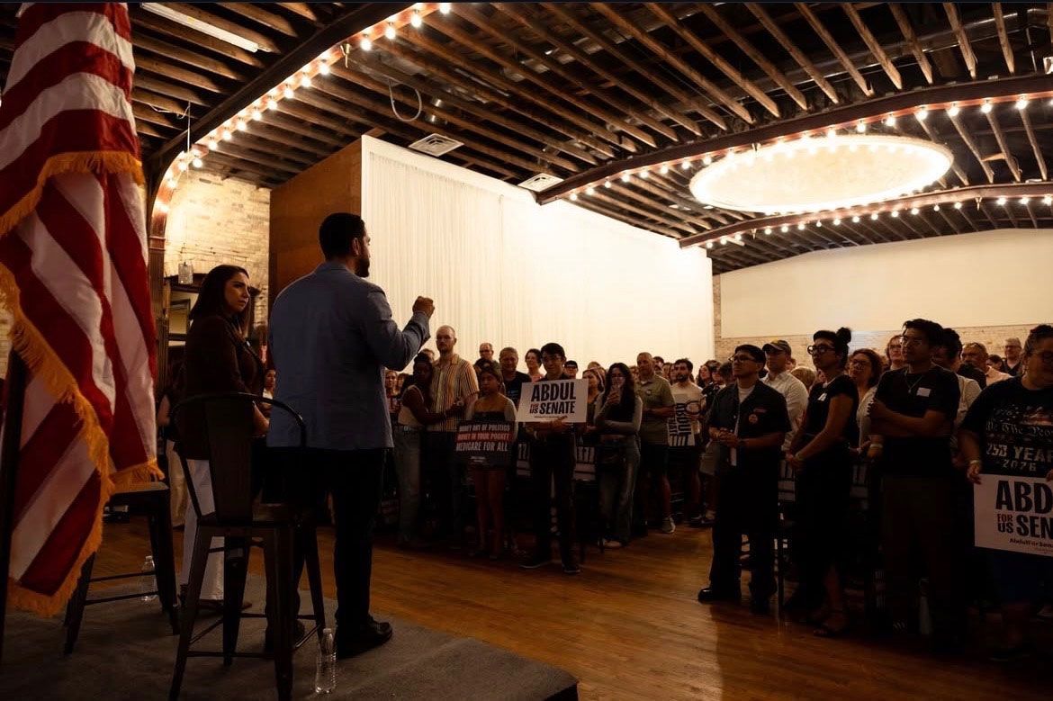 Speaker addressing a crowd in a hall with an American flag and wooden ceiling beams