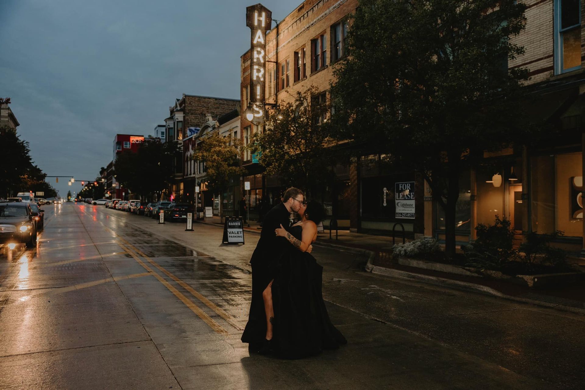 Couple embracing in black on a wet city street at dusk, with storefronts and glowing lights.