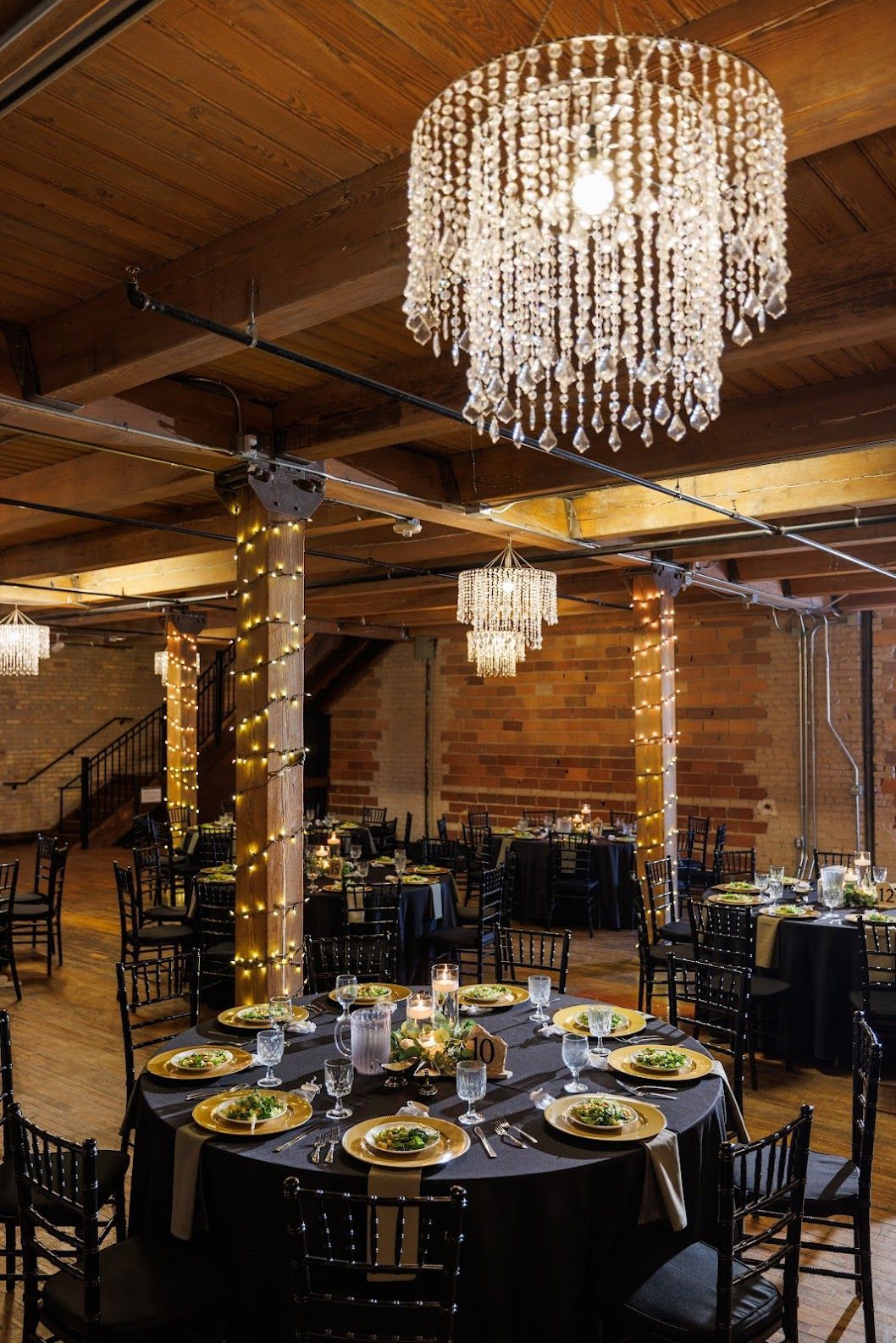 Elegant rustic banquet hall with black tables, string lights, and a crystal chandelier under a wooden ceiling