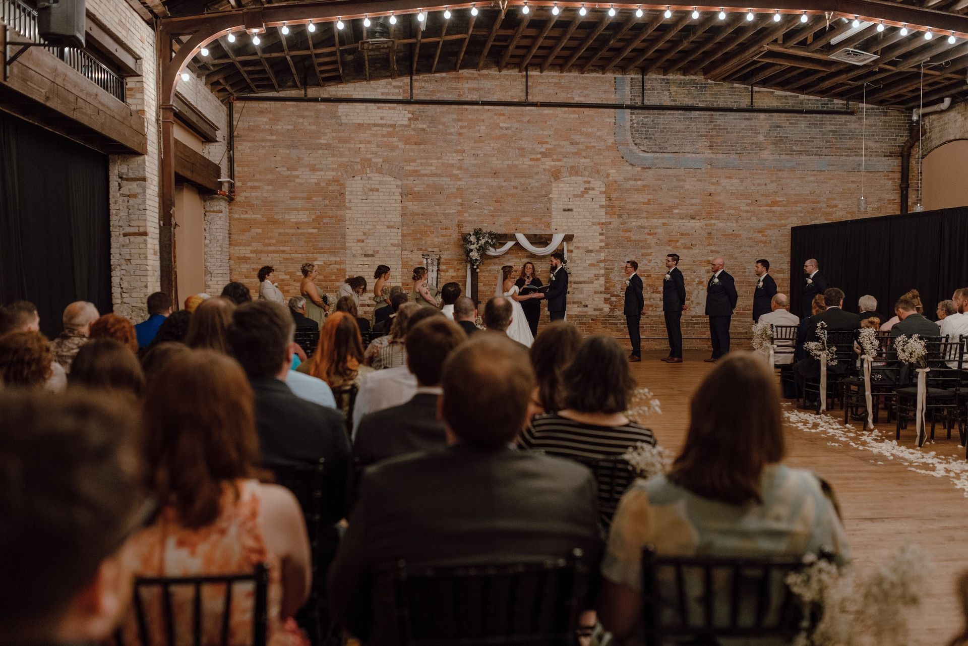 Guests seated in a rustic hall watch a wedding ceremony under warm string lights