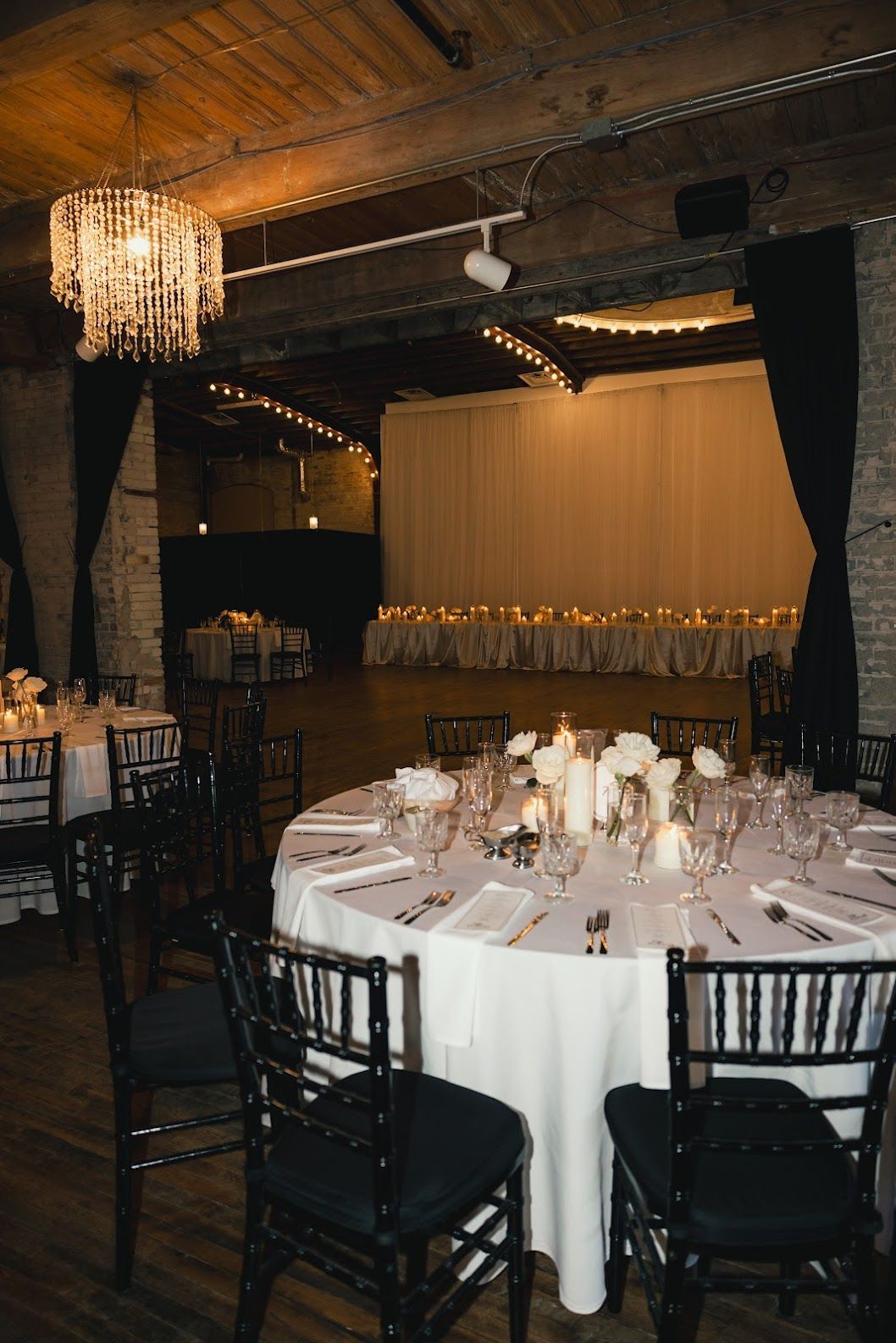 Elegant banquet hall with round white-clothed tables, black chairs, chandeliers, and a lit stage