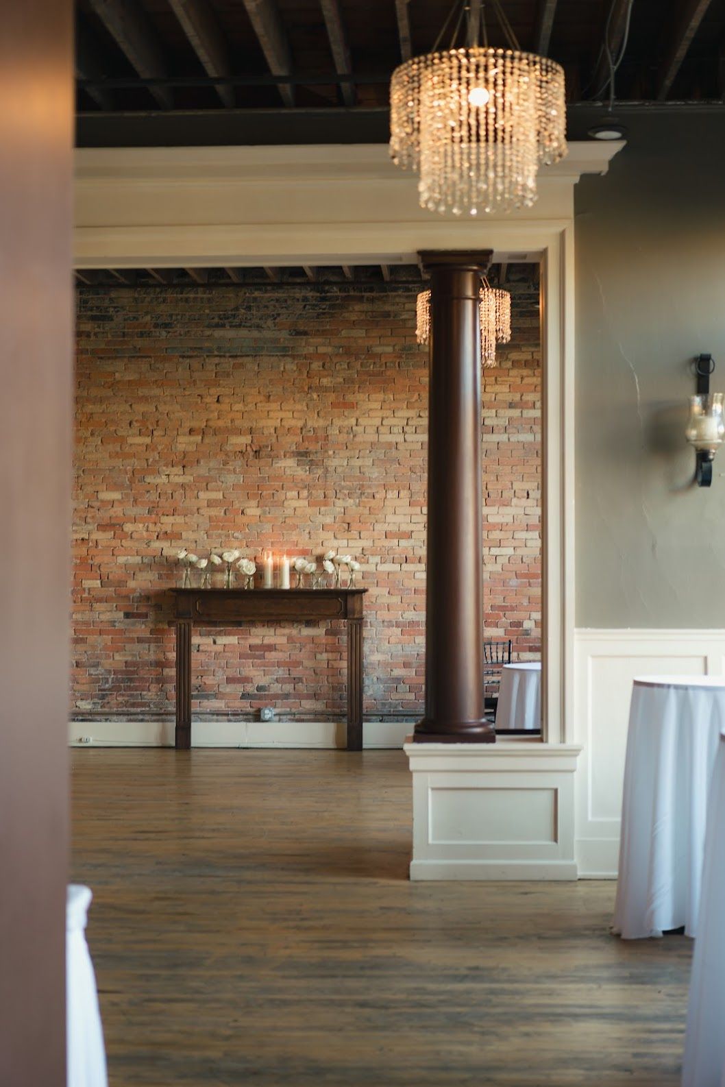 Elegant brick-walled room with wood floors, dark beams, chandelier, and a small table against the back wall