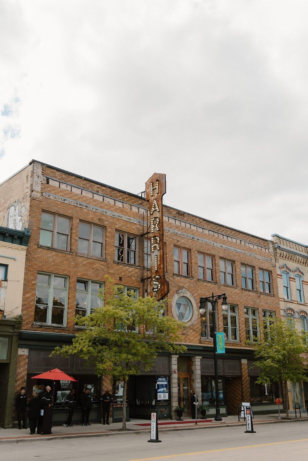 Brick building with large windows, trees, and people outside under an overcast sky.