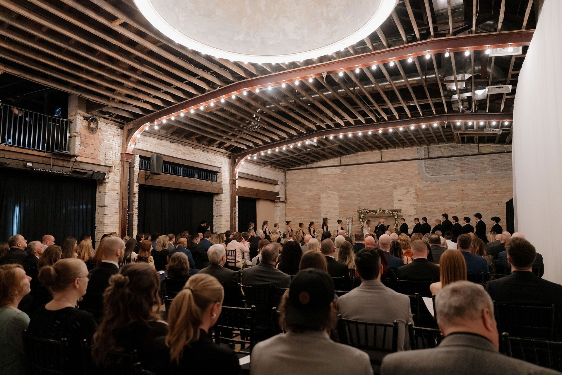 Audience seated in a modern auditorium, facing a stage under curved wooden ceiling beams